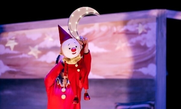 A clown puppet is held above someone wearing red, holding a model of the moon