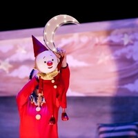 A clown puppet is held above someone wearing red, holding a model of the moon