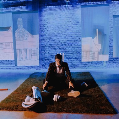 A young Asian man sits on an astro-turf  surrounding by cricket pads and bat with a blue wall and projection behind him