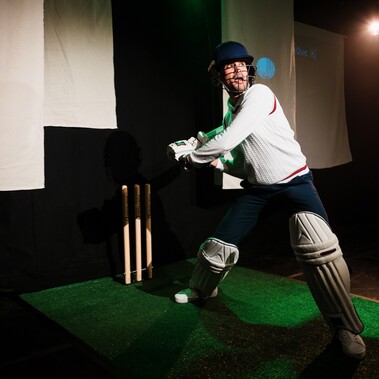 An Asian man dressed in cricket whites swings the bat behind him. He is standing on astro-turf with cricket stumps and white banners theatre spotlights behind him.