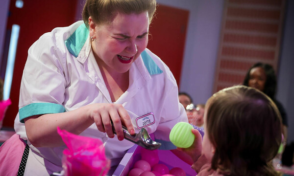 Actor dressed in 1950s style diner pink costume serves a yellow ball to a toddler