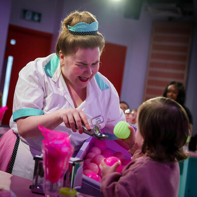 Actor dressed in 1950s style diner pink costume serves a yellow ball to a toddler