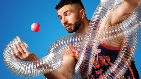 A man wrapped in a clear plastic tube gazes at a juggling ball aimed for the tube