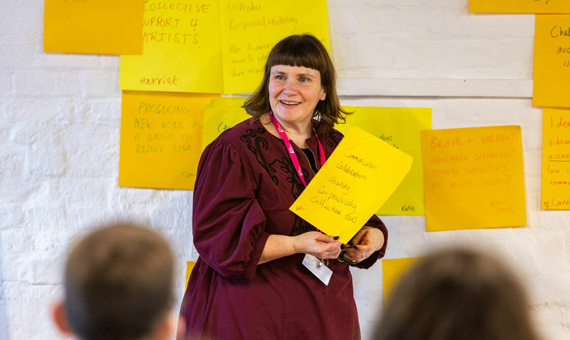 A woman with mid brown hair, a pink lanyard and deep red dress holds a large post it, she's smiling and facing other people