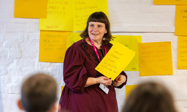 A woman with mid brown hair, a pink lanyard and deep red dress holds a large post it, she's smiling and facing other people