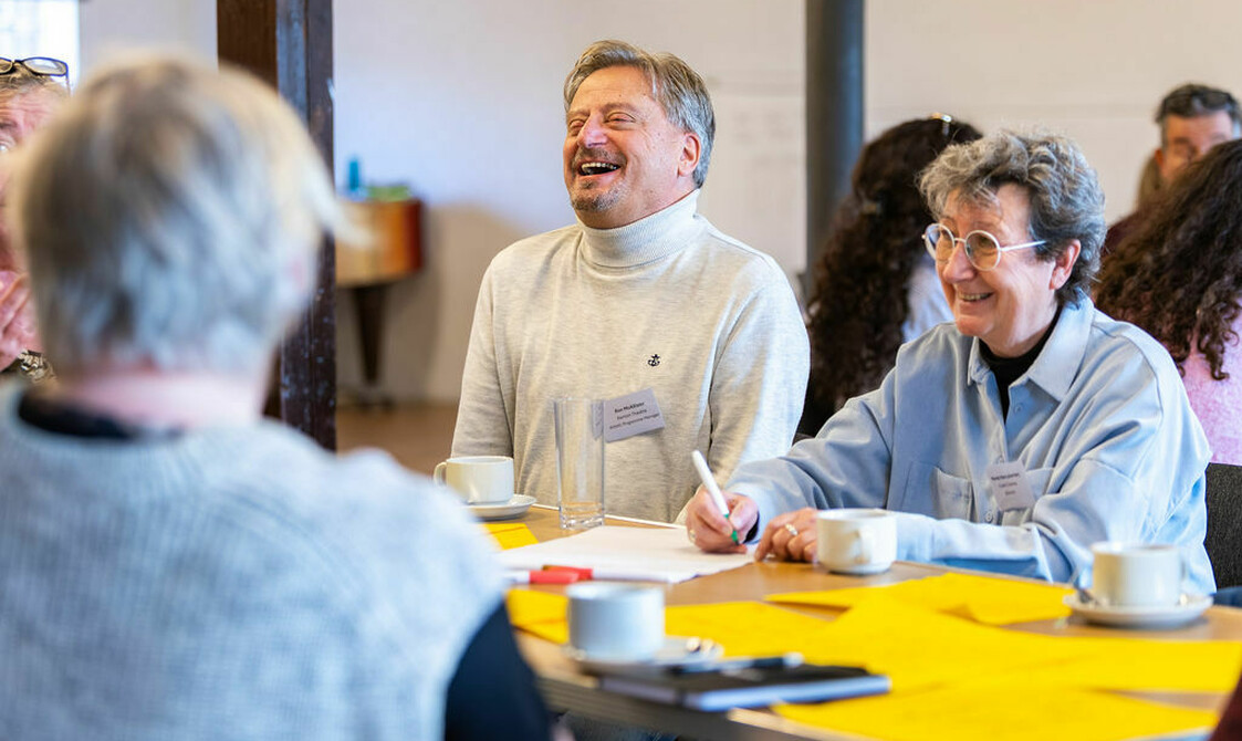 Man in white turtle neck laughing sat next to woman in blue shirt holding a pen, smiling. house Network Day 2026