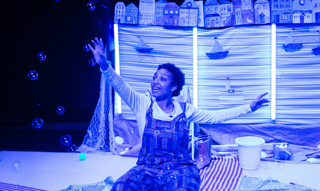 Performer sat on a box drenched in blue lighting - she is reaching for bubbles.