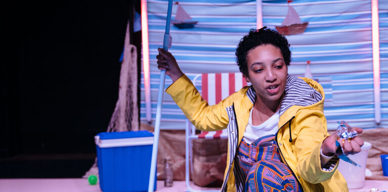 Performer kneeled down in yellow raincoat holding red-striped beach umbrella.