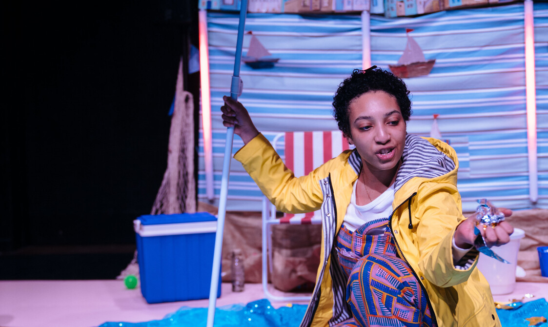 Performer kneeled down in yellow raincoat holding red-striped beach umbrella.