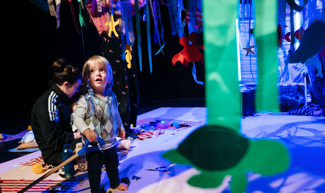 A toddler in a vest sweater stands amongst the blue set during Stay and Play. An adult in a black top sits behind them.