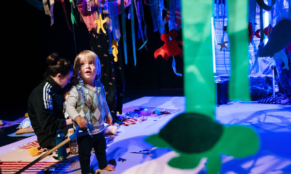 A toddler in a vest sweater stands amongst the blue set during Stay and Play. An adult in a black top sits behind them.