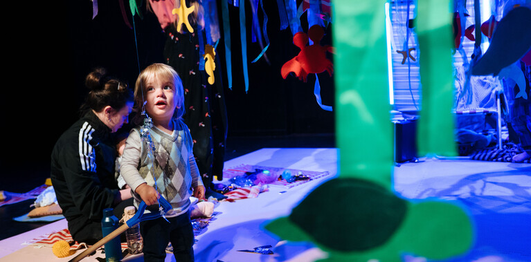 A toddler in a vest sweater stands amongst the blue set during Stay and Play. An adult in a black top sits behind them.