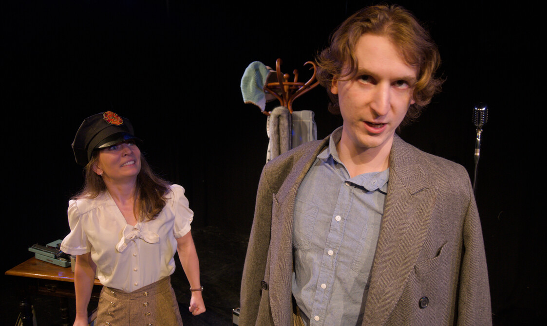 Male performer stands in the foreground looking directly at the camera while the other performer has a hat on grinning at him intensely.