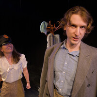 Male performer stands in the foreground looking directly at the camera while the other performer has a hat on grinning at him intensely.