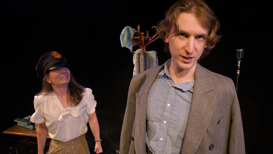 Male performer stands in the foreground looking directly at the camera while the other performer has a hat on grinning at him intensely.