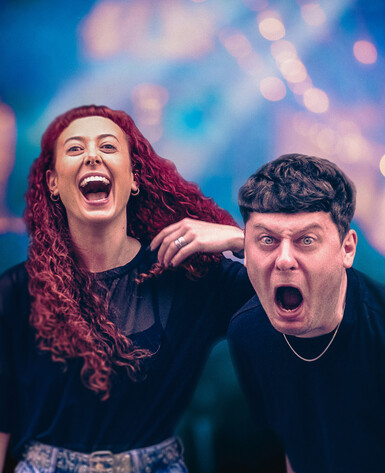 Actor playing Sarah is smiling and looking out past the camera. Actor playing Jamie is mid-shouting intensely into the camera.