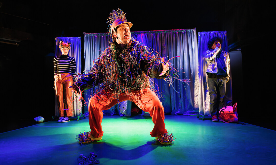 Performer dressed as a fox and a performer as the secret agent are background looking scared. One performer, dressed in electrical wires, hangs out their tongue in the foreground.