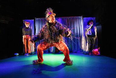 Performer dressed as a fox and a performer as the secret agent are background looking scared. One performer, dressed in electrical wires, hangs out their tongue in the foreground.