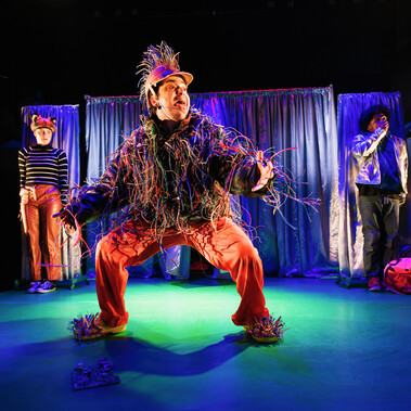 Performer dressed as a fox and a performer as the secret agent are background looking scared. One performer, dressed in electrical wires, hangs out their tongue in the foreground.
