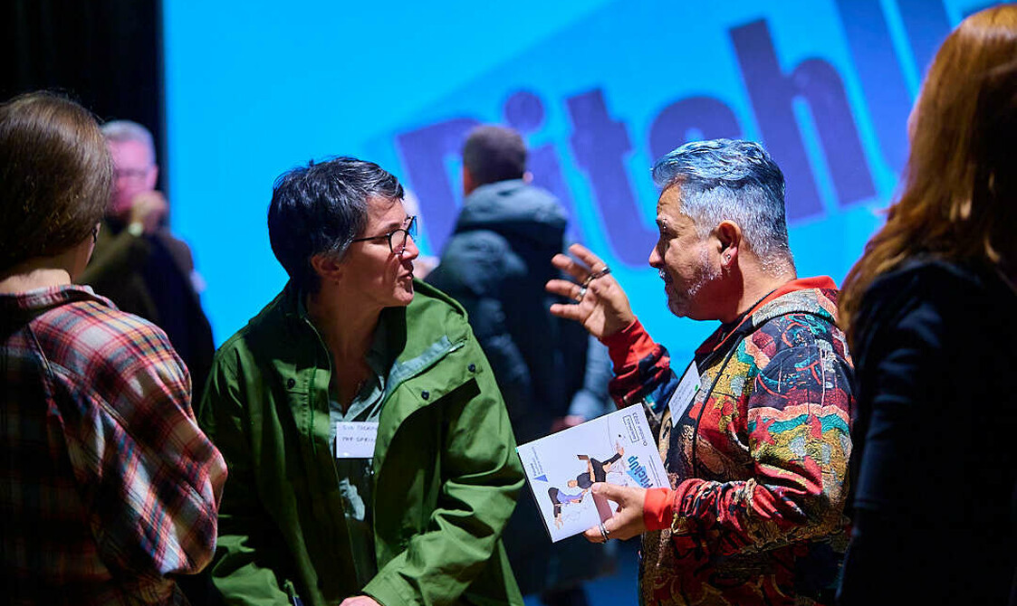 Group of people mid conversation at an event, animated! Pitch Up on the screen behind them.