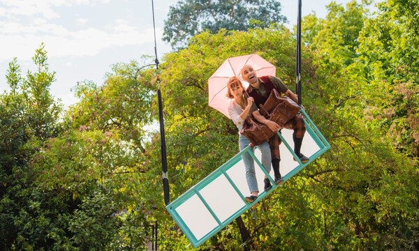 A rig hangs tilted from the sky. On it a white woman holds a pink umbrella and looks worried. A white man smiles with his arm wrapped around a big brown suitcase.