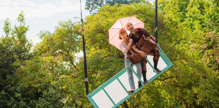 A rig hangs tilted from the sky. On it a white woman holds a pink umbrella and looks worried. A white man smiles with his arm wrapped around a big brown suitcase.