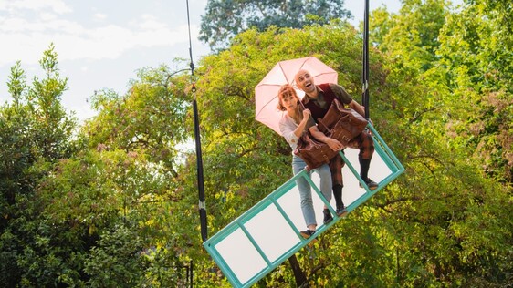 A rig hangs tilted from the sky. On it a white woman holds a pink umbrella and looks worried. A white man smiles with his arm wrapped around a big brown suitcase.
