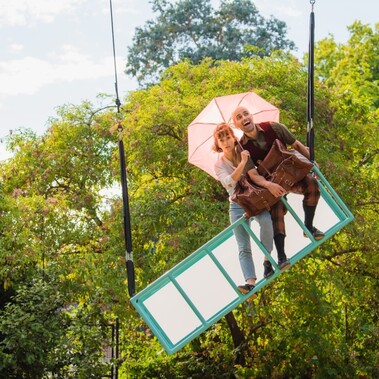 A rig hangs tilted from the sky. On it a white woman holds a pink umbrella and looks worried. A white man smiles with his arm wrapped around a big brown suitcase.