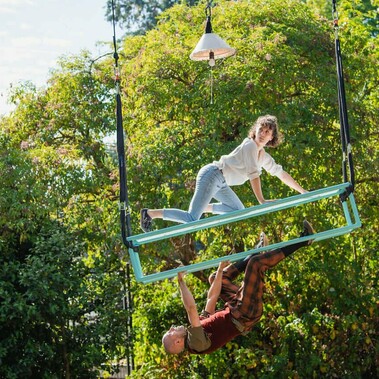 A blue rig hangs from the sky. A lampshade dangles over a white woman with mid length hair. A white man hangs under the rig by his hands, his feet hooked on.