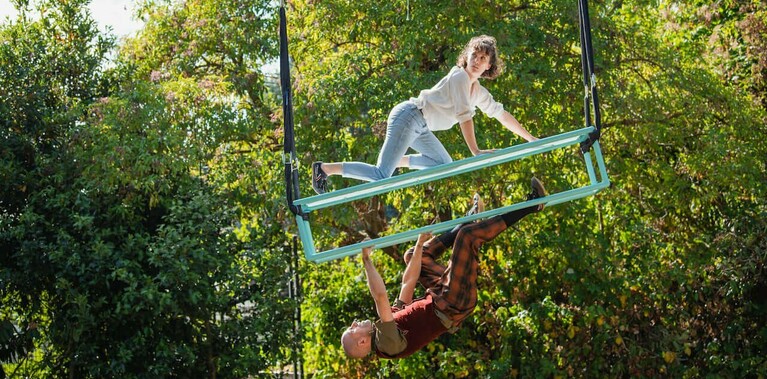 A blue rig hangs from the sky. A lampshade dangles over a white woman with mid length hair. A white man hangs under the rig by his hands, his feet hooked on.