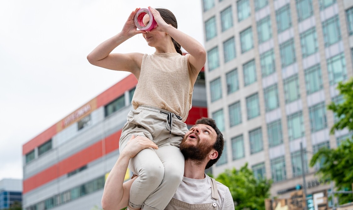 Two white people with brown hair on a bridge, both wearing yellow tops. The woman is standing on the man's head reaching up to the sky with one hand. He has tape wrapped around his chest and mouth.