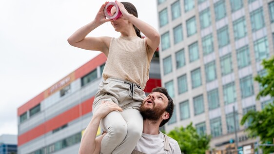 Two white people with brown hair on a bridge, both wearing yellow tops. The woman is standing on the man's head reaching up to the sky with one hand. He has tape wrapped around his chest and mouth.