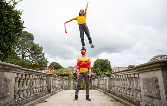Two white people with brown hair on a bridge, both wearing yellow tops. The woman is standing on the man's head reaching up to the sky with one hand. He has tape wrapped around his chest and mouth.