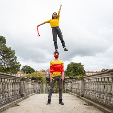 Two white people with brown hair on a bridge, both wearing yellow tops. The woman is standing on the man's head reaching up to the sky with one hand. He has tape wrapped around his chest and mouth.