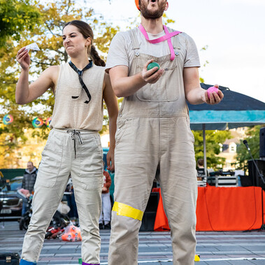 Two white people outside, a tree and marquee in background. Both pulling faces and have tape on their legs. He is juggling and has green tape on his head.