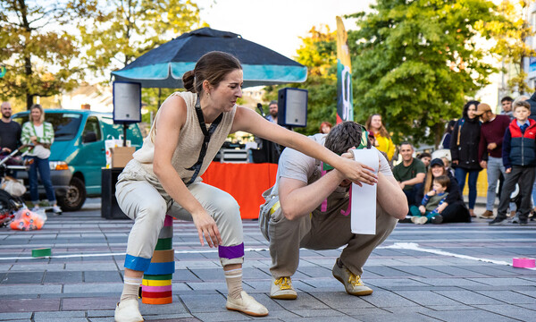 Outside paved area with trees and marquee in background. A white woman and man crouch, her arm across him, his face is covered by a toilet roll.