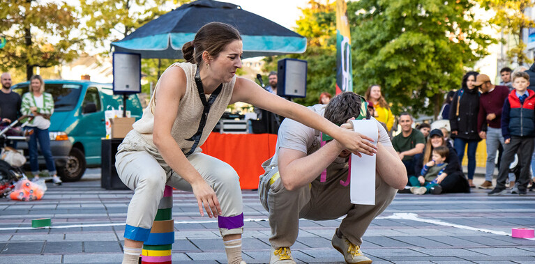Outside paved area with trees and marquee in background. A white woman and man crouch, her arm across him, his face is covered by a toilet roll.