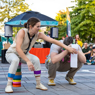 Outside paved area with trees and marquee in background. A white woman and man crouch, her arm across him, his face is covered by a toilet roll.