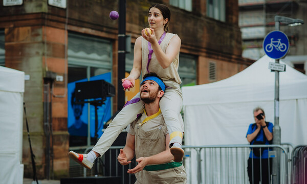 Two people in front of buildings and white tents, both wearing cream clothing. The woman is sitting around the man's neck, they are both juggling balls.