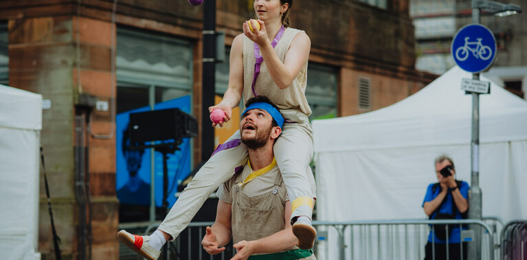 Two people in front of buildings and white tents, both wearing cream clothing. The woman is sitting around the man's neck, they are both juggling balls.
