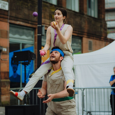 Two people in front of buildings and white tents, both wearing cream clothing. The woman is sitting around the man's neck, they are both juggling balls.
