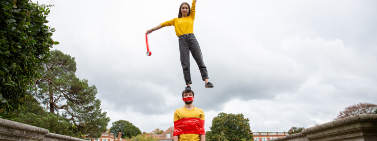 Two white people in yellow tops on a bridge. The woman is stood on the man's head reaching up with one arm. He has tape wrapped around his chest and mouth.
