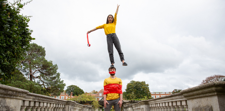 Two white people in yellow tops on a bridge. The woman is stood on the man's head reaching up with one arm. He has tape wrapped around his chest and mouth.
