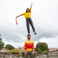 Two white people in yellow tops on a bridge. The woman is stood on the man's head reaching up with one arm. He has tape wrapped around his chest and mouth.