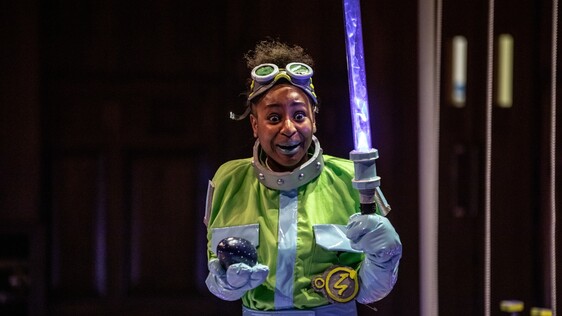 Woman wearing goggles on her forehead, a yellow cape holding a light sabre