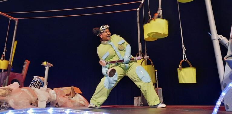 Woman in yellow boiler-suit surrounded by hanging yellow buckets and neon lights on a theatre stage