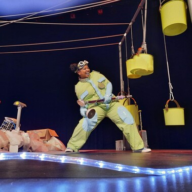 Woman in yellow boiler-suit surrounded by hanging yellow buckets and neon lights on a theatre stage