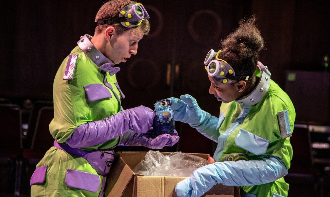 Two actors in yellow outfits and goggles on their heads and long gloves hold a one eyed puppet over a box