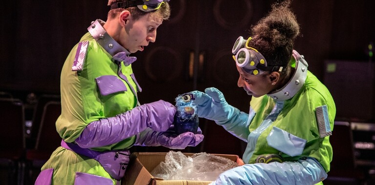 Two actors in yellow outfits and goggles on their heads and long gloves hold a one eyed puppet over a box