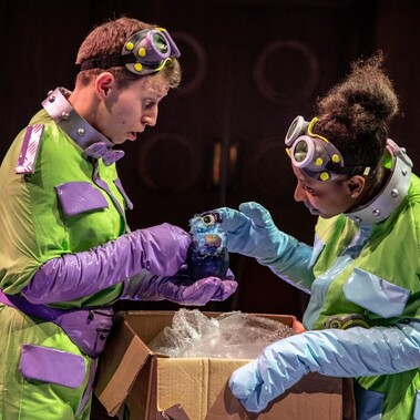 Two actors in yellow outfits and goggles on their heads and long gloves hold a one eyed puppet over a box